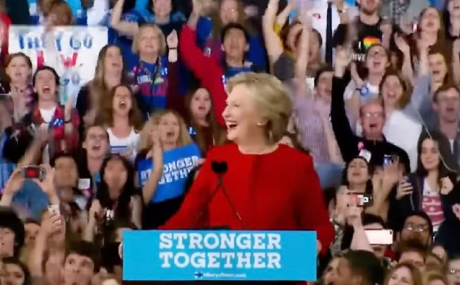 A person in a red outfit stands at a podium with a "Stronger Together" sign, surrounded by a cheering crowd holding signs.